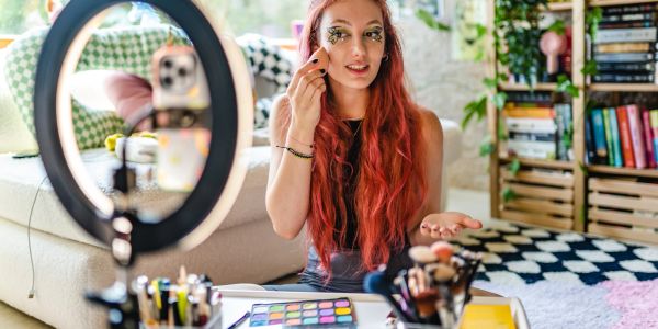 Young woman applying makeup in front of a ring light and camera.
