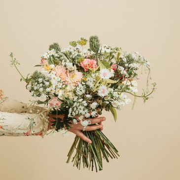 Hand holding a delicate bouquet of mixed wildflowers against a beige background.