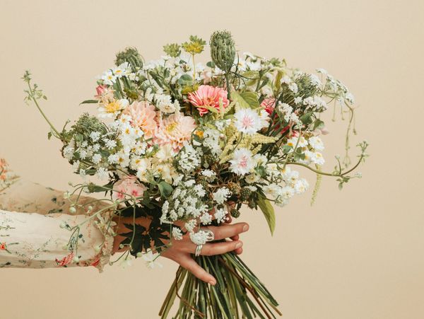 Hand holding a bouquet of wildflowers and greenery against beige background.