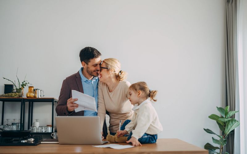 Loving couple standing next to the table smiling and hugging. He is holding a piece of paper with a monthly bill. She is leaning towards him and smiling. Their daughter is sitting on the table looking at the laptop computer.
