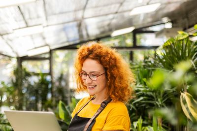 A woman with curly red hair working on a laptop in a greenhouse.