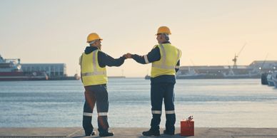 Two construction workers in safety gear fist bump by the waterfront.