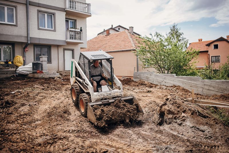 Construction site worker driving a small excavator loading and carrying dirt at the back yard construction site.