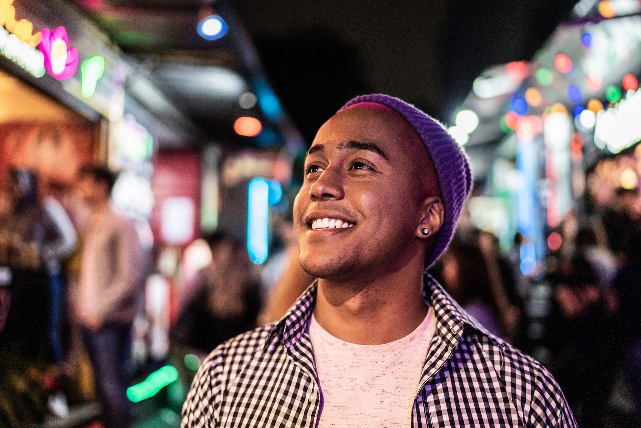 Smiling man with pink hair and purple beanie in a lively, colorful street at night.