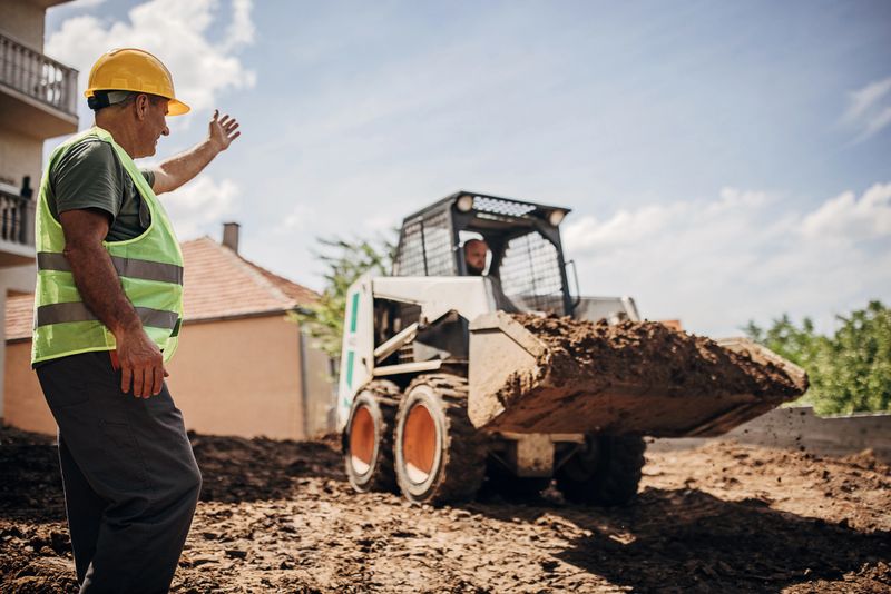 Mature male engineer coordinating worker who is working with small excavator moving dirt at construction site.