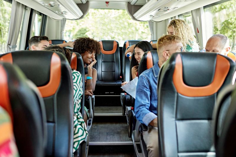 View from front of motor coach as diverse group of relaxed tourists smile and converse during late spring road trip.