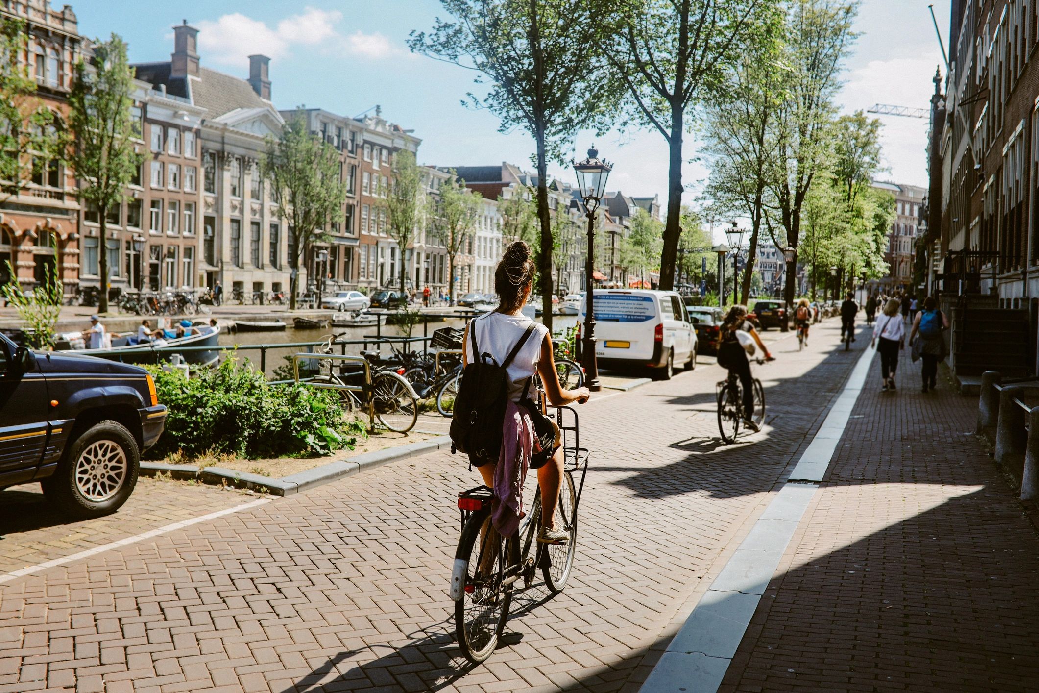 Cyclists ride along a sunny canal-side street lined with parked cars, trees, and historic buildings.