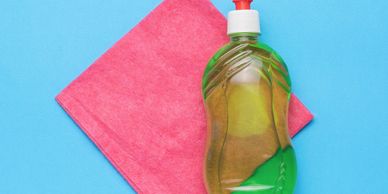 Green dish soap bottle and pink cleaning cloth on blue background.