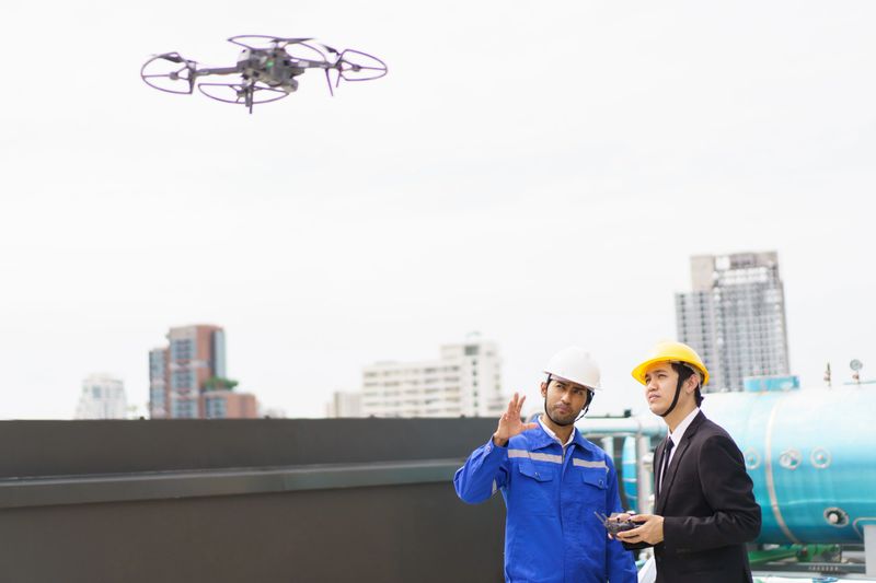 Professional Asian civil engineers using the unmanned aerial vehicle or UAV - drone for inspecting the building structure, 2 Asian foreman flying a drone to the building roof top area.