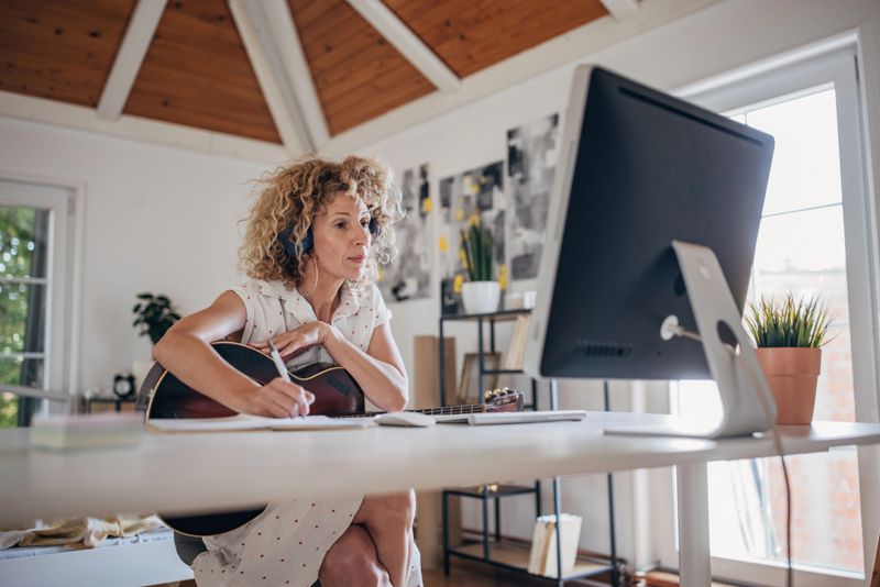 One woman, mature female with headphones sitting at the desk in bedroom at home, she is holding acoustic guitar while writing notes for online course.