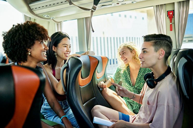 Diverse group of casually dressed women in late teens and 20s sitting face to face and laughing as they wait for motor coach to depart on vacation road trip.