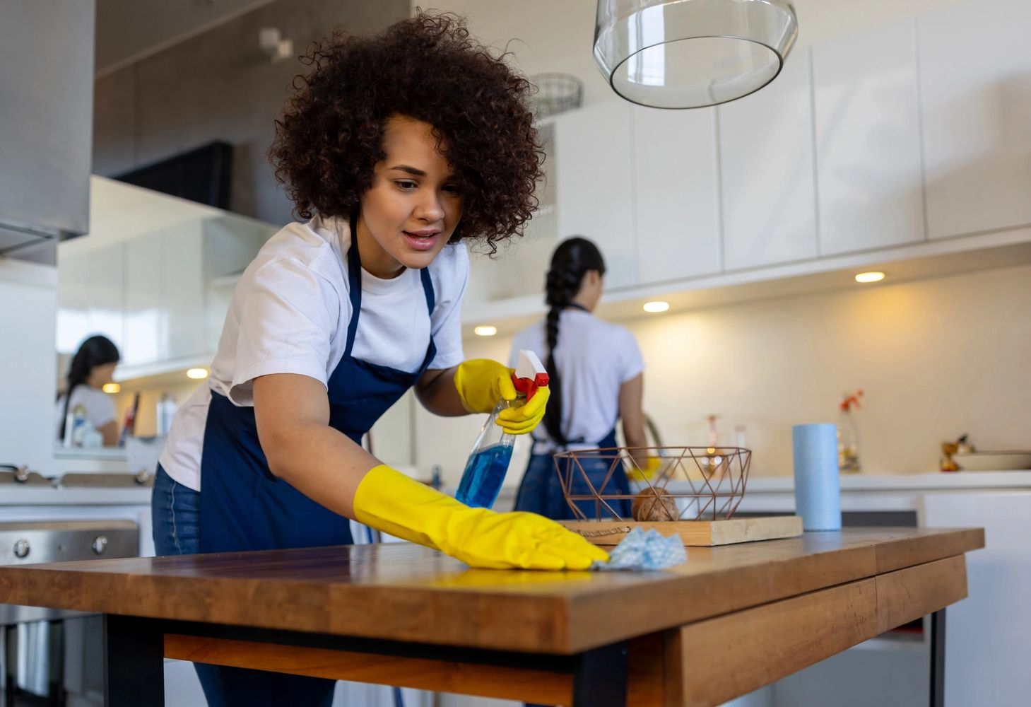 Two women cleaning a kitchen, one wiping a wooden table.