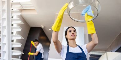 Woman cleaning a light fixture with gloves, man mopping floor in background.