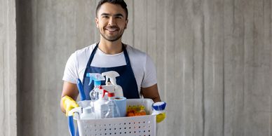 Smiling man in an apron holding a basket of cleaning supplies.