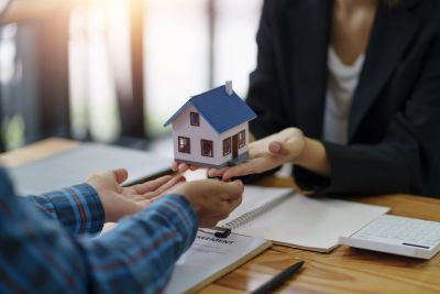 Two people exchanging a miniature house model over a contract on a wooden table.