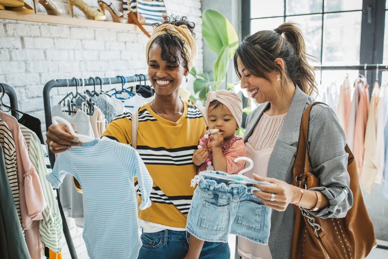 Young female gay couple at thrift store with baby girl, talking and choosing clothes.