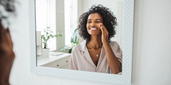 Woman smiling while applying skincare in front of a mirror.