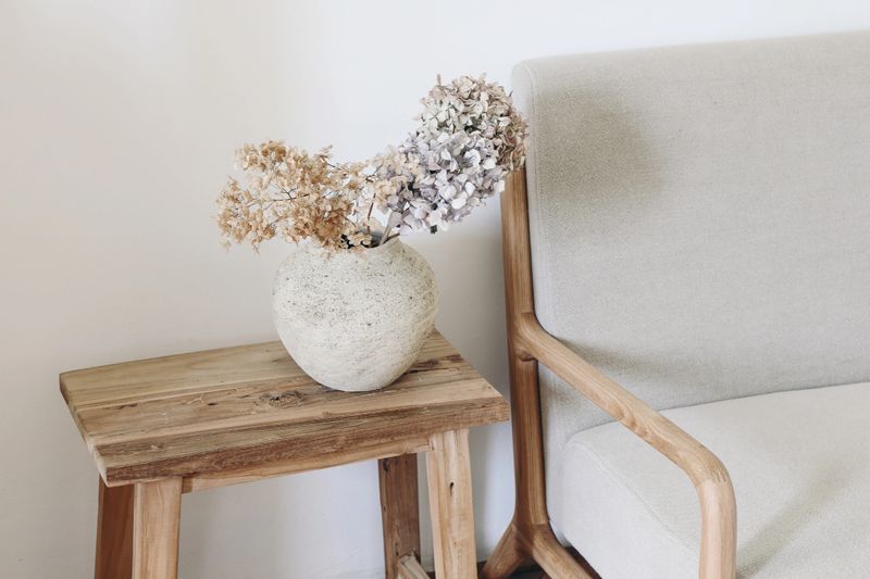 Fall still life photo. Rustic textured vase with dry hydrangea flowers on old wooden bench. Blurred linen mid century sofa background. White wall. Scandinavian interior, boho elegant home decor.