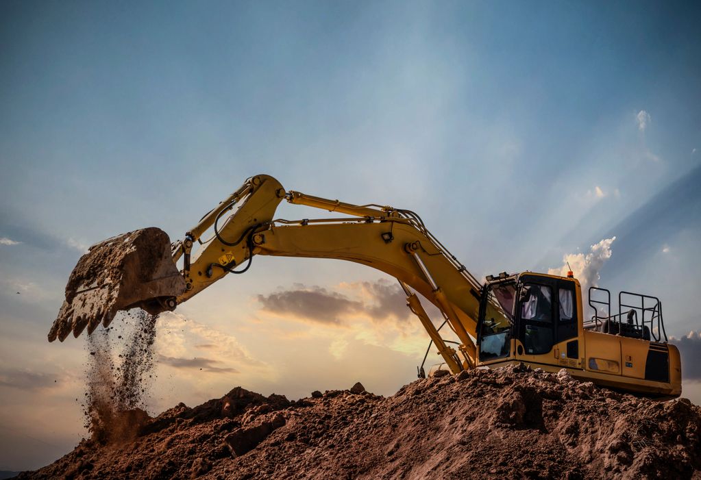 Yellow excavator digging and dumping soil at a construction site during sunset.
