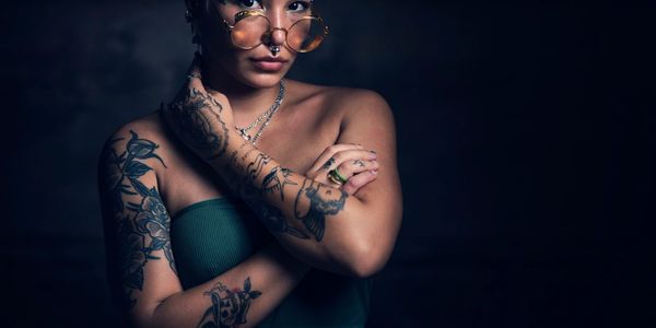 Woman with tattoos and glasses poses against dark background.