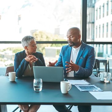 Two professionals discussing work at a conference table with a laptop.