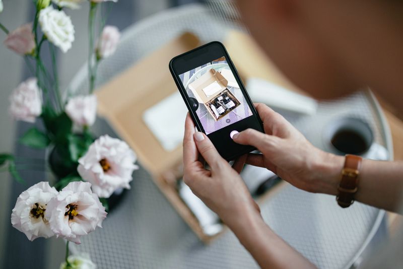 An unrecognizable woman holding a phone and taking a photo of the carton gift box laid on the table in front of her.