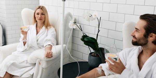 Couple in bathrobes receiving IV therapy while relaxing with drinks in a spa-like setting.