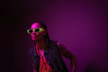 Person with neon green sunglasses and studded vest under purple lighting.