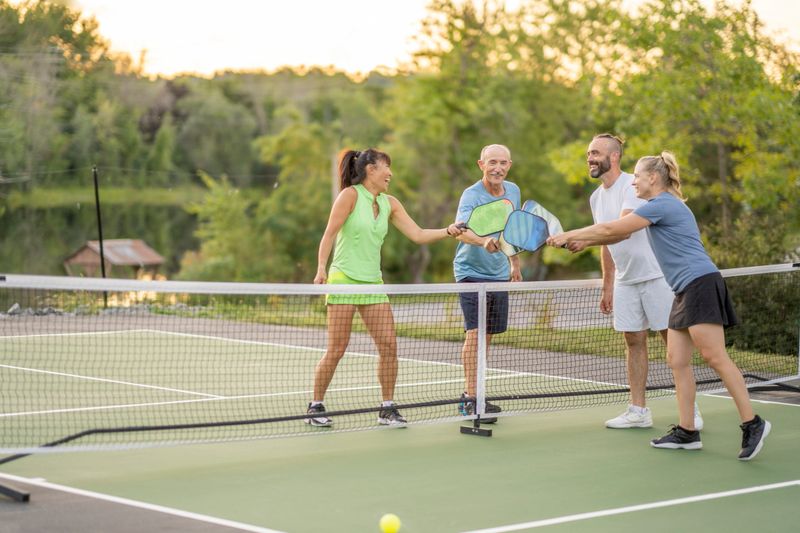 A group of four adults are seen tapping racquets to show sportsmanship after a Pickleball game together.  They are each dressed comfortably in athletic wear and are smiling as they enjoy their time in the fresh air.