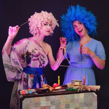 Two women in colorful wigs play musical triangles with various instruments on a table.