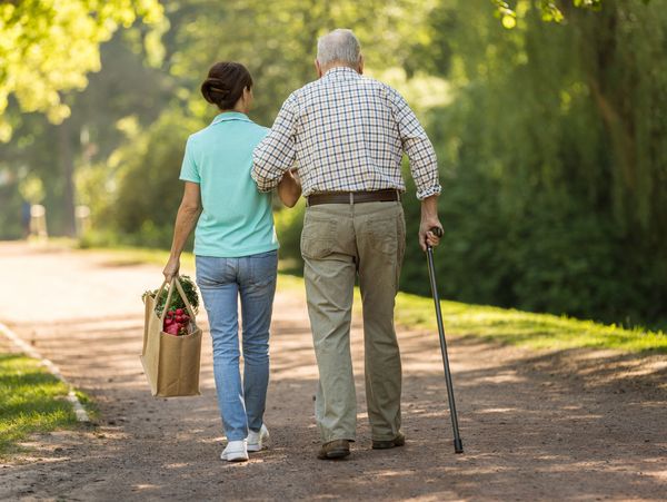 Young woman helps elderly man with cane walk outdoors.