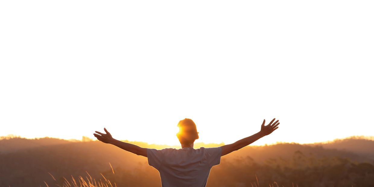 Person with arms outstretched facing a bright sunrise over hills.