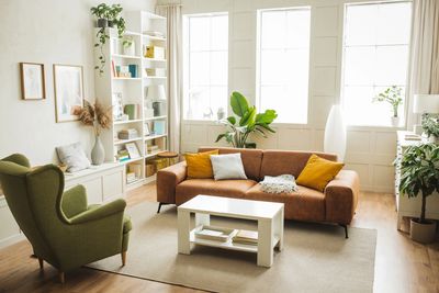 Bright living room with brown sofa, green armchair, plants, and white bookshelf.