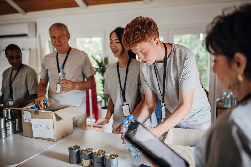 Diverse group of male and female volunteers sorting donated canned food and water bottles into cardboard boxes in charity center.