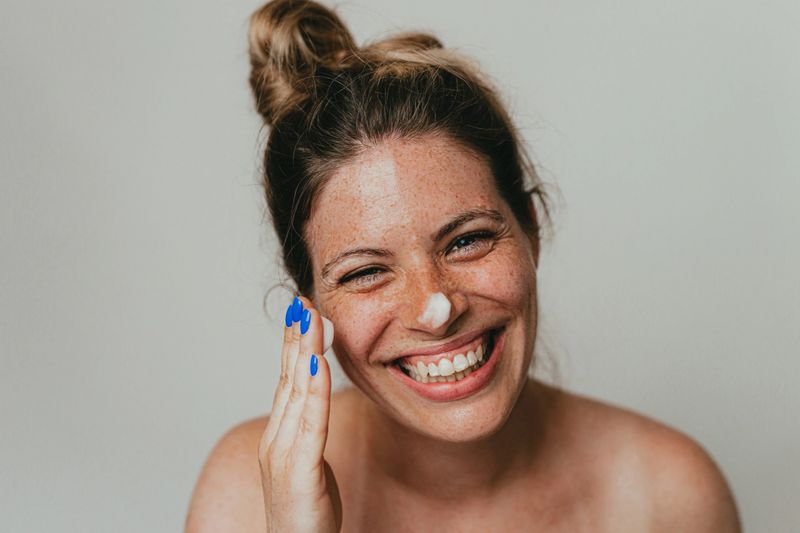 Portrait of a beautiful young woman with freckles applying a face cream, studio shot in front of white background
