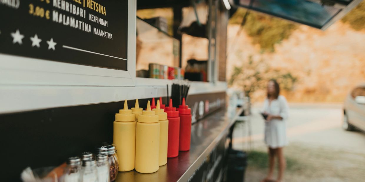 Condiment bottles lined up on a food truck counter with a blurred person in the background.