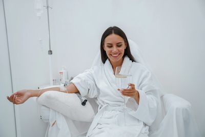 A woman in a white robe receiving IV therapy while holding a glass of water with lemon.