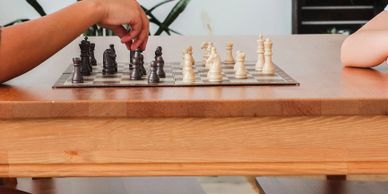 Two people playing chess on a wooden table, one about to move a black piece.