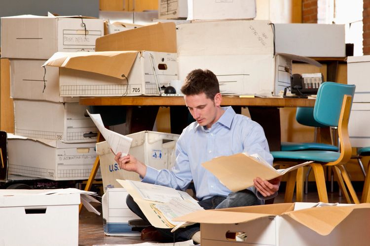 Man sorting through paperwork surrounded by boxes in an office.