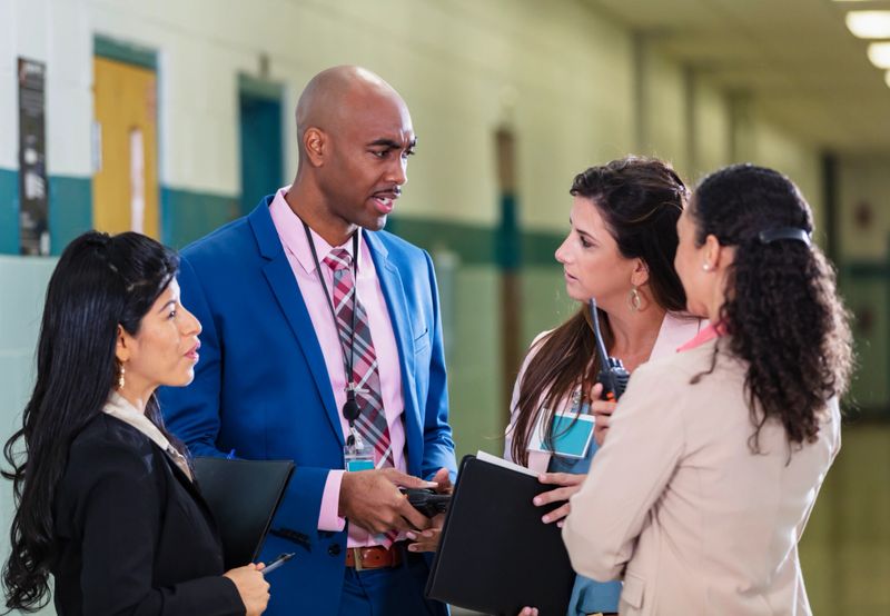 A multiracial group of four teachers or school administrators standing in circle in a school hallway having a meeting. The African-American man wearing a full suit perhaps is the school principal.