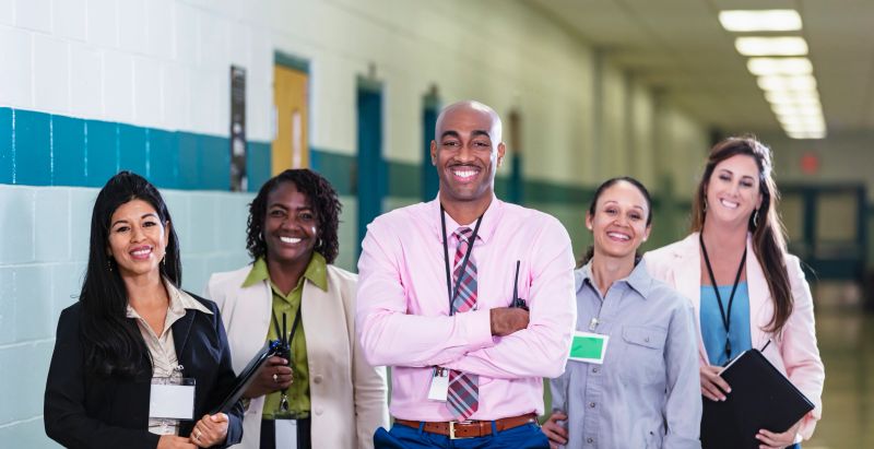 A multiracial group of five teachers or school administrators standing together in a school hallway smiling confidently at the camera in a group portrait. The African-American man in the middle is the team leader, perhaps the school principal. He is the only male in the group. The focus is on him and the Hispanic woman standing on the left.