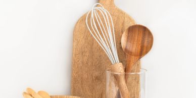 Wooden kitchen utensils and whisk in a glass with a wooden cutting board.