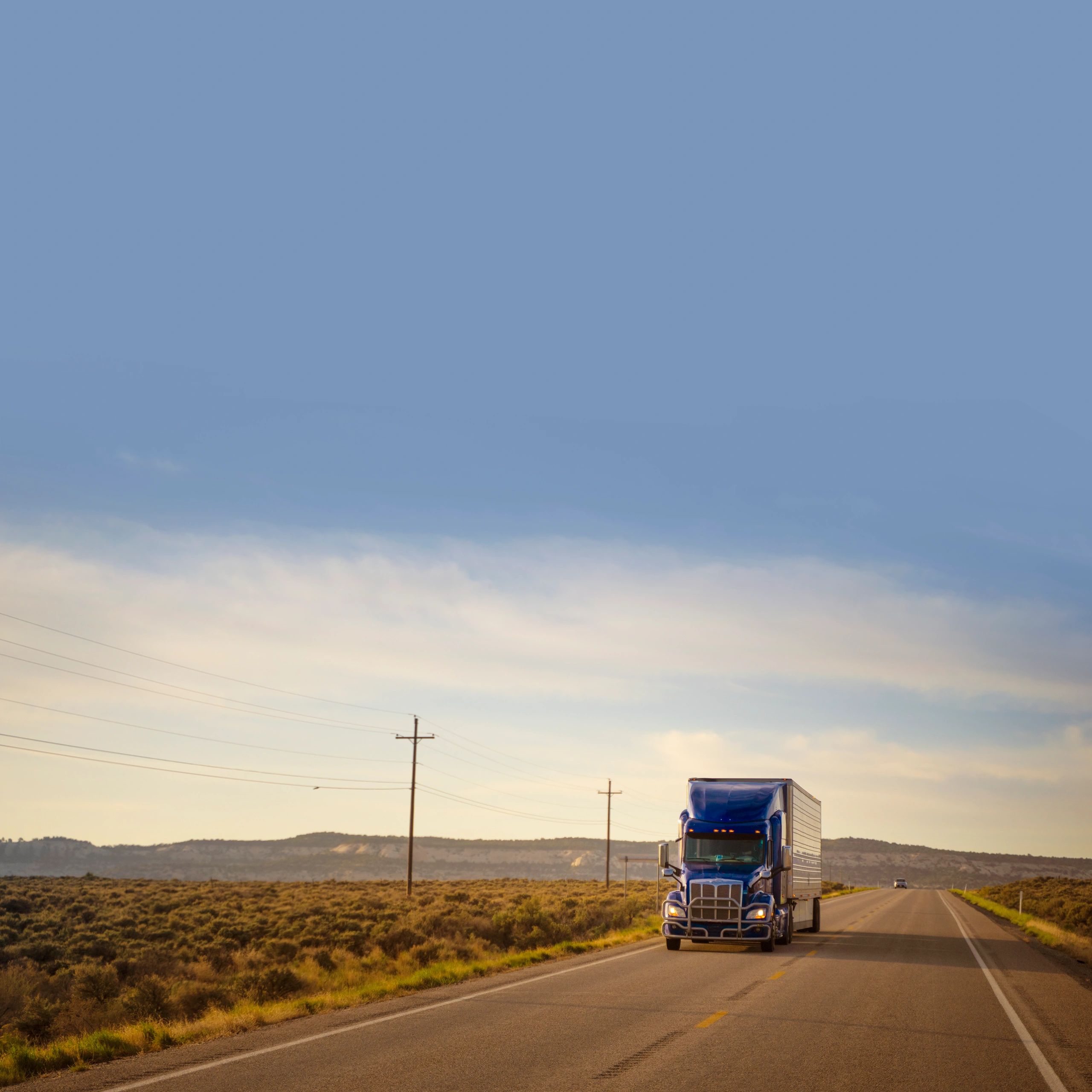 Blue semi truck driving on an open highway at sunset.