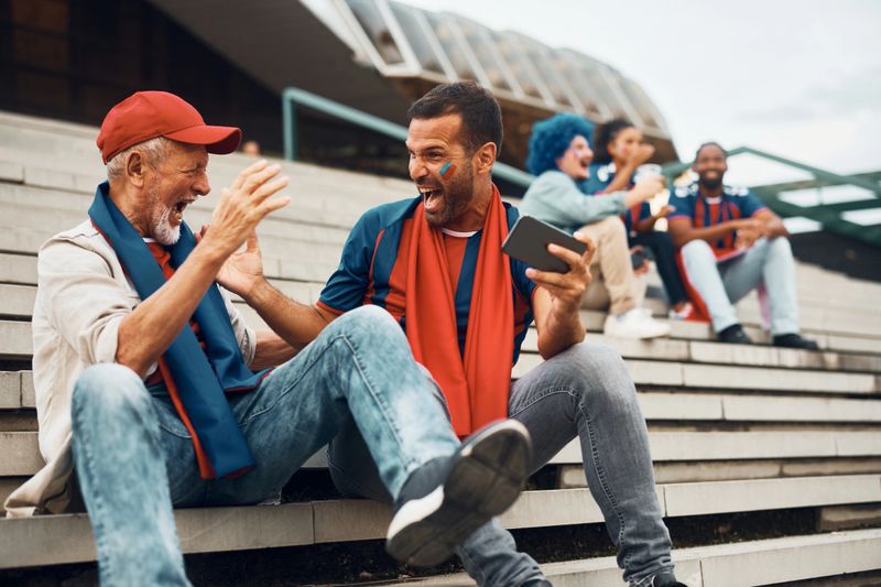 Cheerful sports fan and his senior father celebrating while watching soccer match on smart phone outdoors.