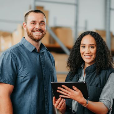 Two colleagues smiling with a tablet in a warehouse.