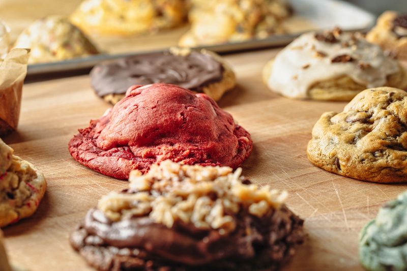 Giant-size homemade cookies on a cutting board showcasing Smores Chocolate, Red Velvet, Chocolate Chip, Sugar Sprinkles and other various styles