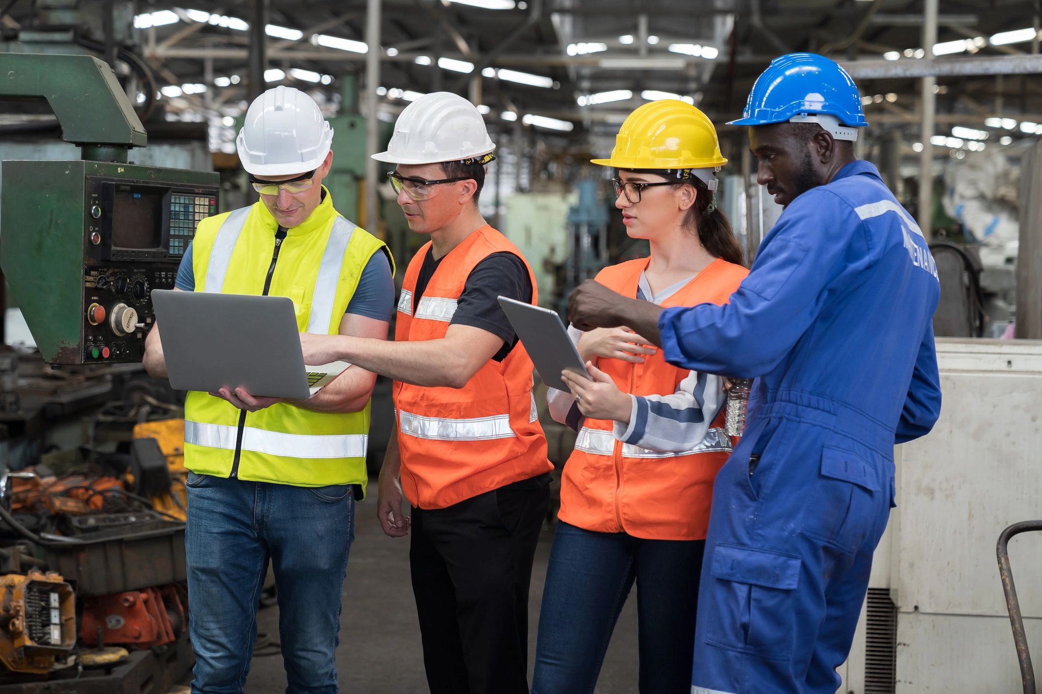 Four factory workers in safety gear discussing work using a laptop and tablet.
