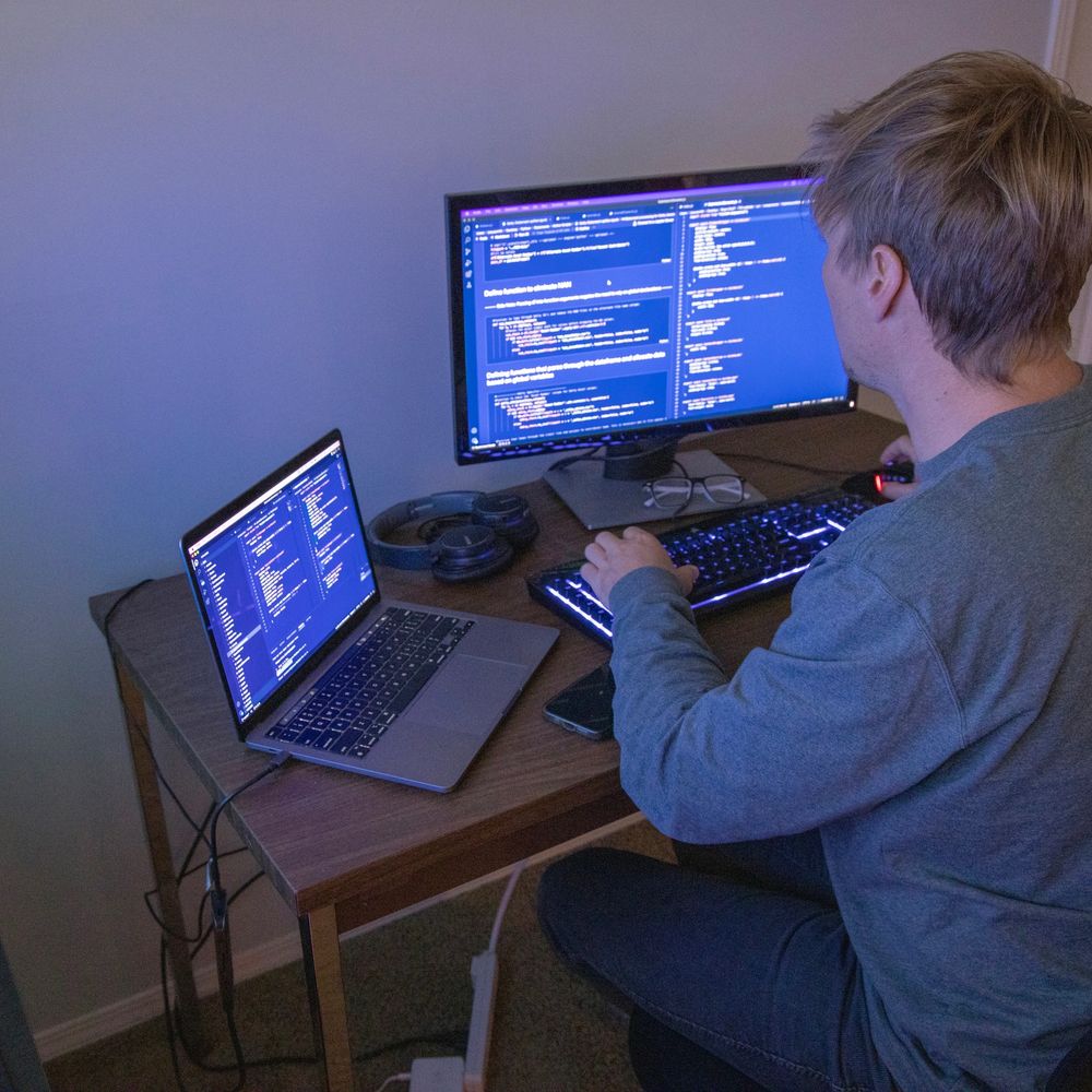 Person coding on a dual-screen computer setup with laptop and monitor.