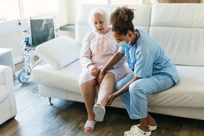A nurse examines an elderly woman's leg in a cozy living room.