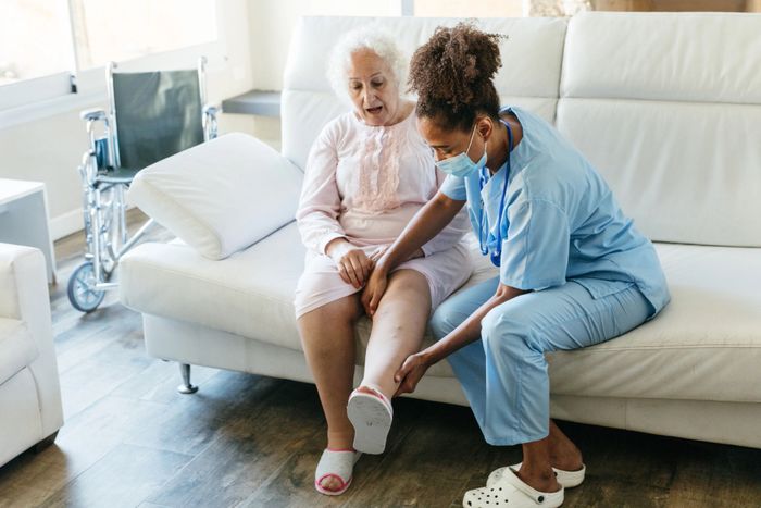 A nurse examines an elderly woman's leg in a cozy living room.